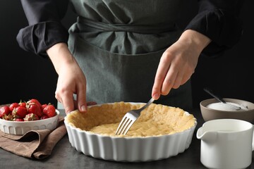 Shortcrust pastry. Woman making holes in raw dough with fork at grey table, closeup