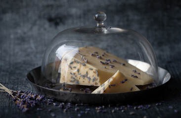 Photo of hard cheese lying under a cheese bell. There is lavender on the cheese. There are also...