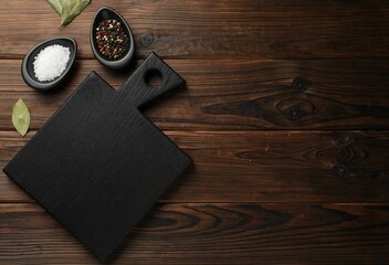 Black cutting board, salt, pepper and bay leaves on wooden table, flat lay. Space for text