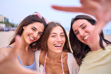 Portrait cheerful group of three attractive European women posing photo doing hand frame gesture on beach. Smiling friends looking through fingers at camera. Gen z joyful female outdoor summer holiday