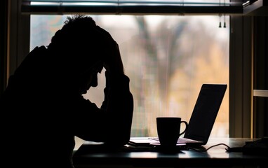 Silhouetted person stressed at desk with laptop and coffee cup by window.