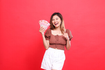 The expression of a cheerful Asian woman holding rupiah money while clenching her fists in front of her, wearing a brown blouse with a red background. for transaction, technology and advertising