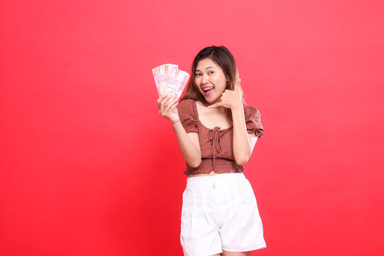 gesture of a young Asian woman, happy and cheerful, holding rupiah money in front of her and a telephone sign. I am wearing a brown blouse with a red background. for transaction and advertising