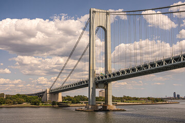 New York, NY, USA - August 1, 2023: N view, Verrazzano-Narrows Bridge E side half and landing at Fort Hamilton. Green coastline with tall buildings on horizon under blue sky