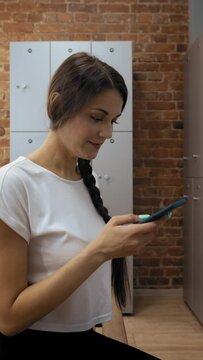 Young woman with long braid closes locker sitting on wooden bench to text with boyfriend on smartphone. Ladies spend time in dressing room before training Vertical Shot.