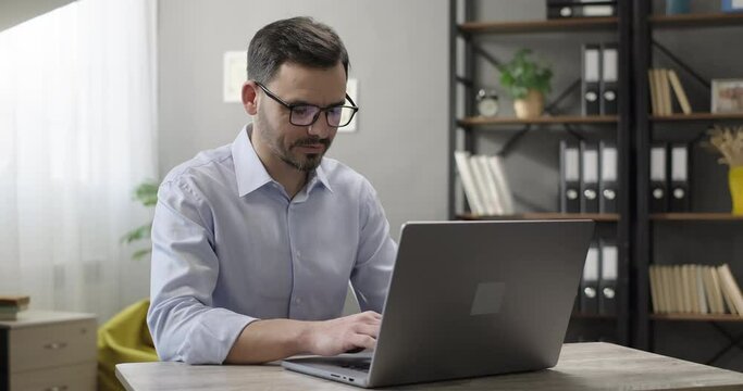 Smiling entrepreneur working on laptop computer at home office. Bearded man typing email, browsing website, analyzing data. Remote work