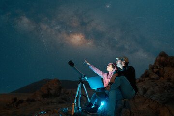 heterosexual couple sitting in the desert watching the stars and Milky Way next to a telescope,...