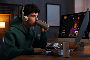 Young man with energy drink and headphones playing video game at wooden desk indoors
