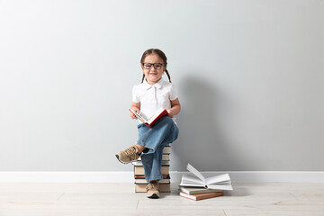 Cute little girl in glasses sitting on stack of books near light grey wall