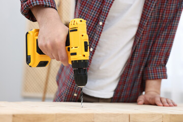 Young handyman working with electric drill at table in workshop, closeup