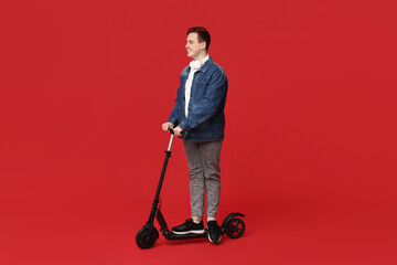 Young man riding modern electric kick scooter with headphones on red background