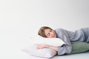 A woman lies comfortably on her side, embracing a white pillow with a tranquil expression. She is wearing a pajamas and resting against a light, solid-colored backdrop, evoking a sense of relaxation