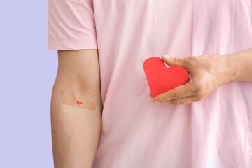 Blood donor with applied medical patch holding paper heart on lilac background, closeup