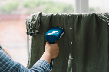 Woman Using a Blue Handheld Steamer on a Green Garment at Home During Daytime. Natural daylight...