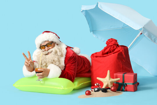 Santa Claus with cocktail and bag of gifts showing victory gesture on blue background. Christmas in July