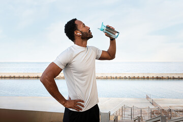 Young Black male runner taking a break to hydrate. African American man drinking water after...