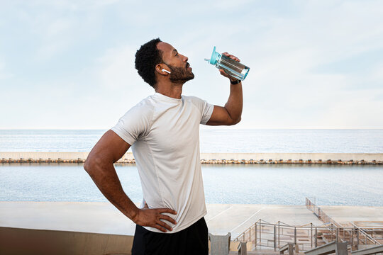 Young Black male runner taking a break to hydrate. African American man drinking water after exercising outdoors.