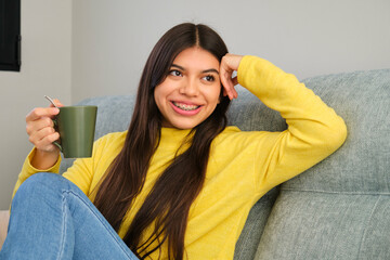 Female smiling teenager with braces and a cup of coffee relaxing on a sofa at home.