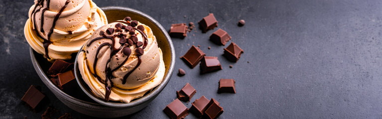 vanilla chocolate ice cream in dark bowl, generously drizzled with liquid chocolate, sprinkled with chocolate chips, broken chocolate bar pieces on dark surface. banner, copy space