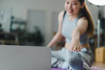 Young Asian woman at home doing exercise in front of open laptop, repeating instructions by...