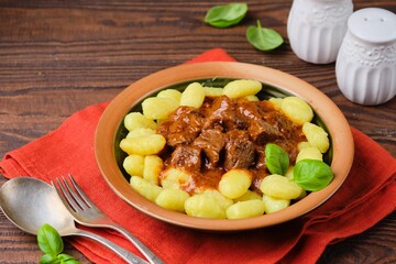 Triestine beef goulash in tomato and sweet paprika sauce, served with potato gnocchi, in a ceramic plate on a brown wooden background. Beef recipes.