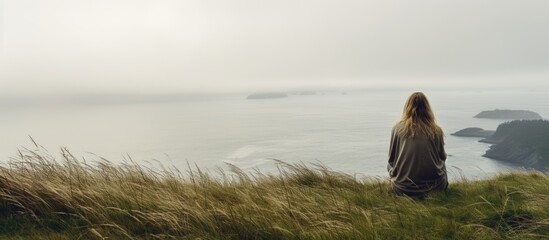 A teenage girl sitting on a grassy hill near the ocean in the Pacific Northwest on a misty day with a background suitable for a copy space image - Powered by Adobe