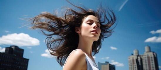 Naklejka premium A beautiful young brunette woman with elegant makeup swaying her head and running her fingers through her hair poses against a backdrop of buildings under a clear blue sky on a sunny day with copy spa
