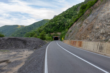  A vehicle enters a tunnel on a well-maintained mountain road with clear skies overhead. The road...