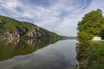  captures a serene river scene with a cabin and boat on the grassy bank, set against a backdrop of cliffs and mountains. The calm water reflects the blue sky and greenery, creating a peaceful 