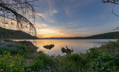 The water reflects the golden hues, while the distant mountains and greenery add depth. The presence of pollution contrasts the natural beauty.