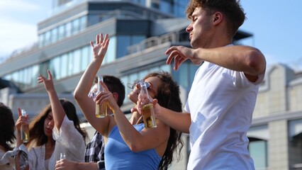 Many young casual people dancing outdoor with drinks at rooftop party on sunny day holding bottles in hands. Happy male and female friends having fun dance at celebration. Close up
