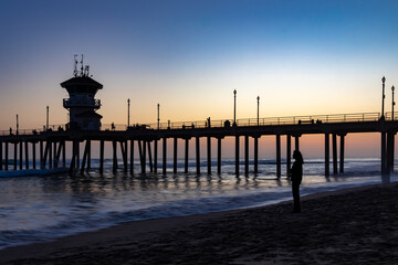 Obraz premium Twilight Sky With Silhouette of Majority of Huntington Beach Pier in Midground and One Person Standing While Looking out to Sea, Distant People in Motion on Pier, California, USA, horizontal 