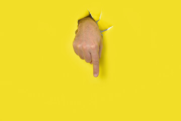 Male hand coming out of a hole in a yellow torn paper background pointing down.