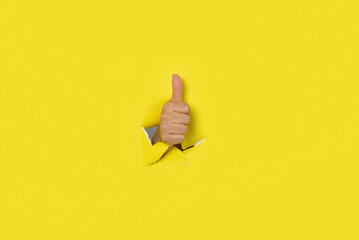 Hand with thumb up as a sign of approval, coming out of the hole of a yellow torn paper background.