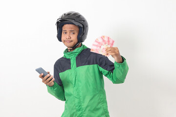 Portrait of Asian online taxi driver wearing green standing against white background, smiling and looking at camera, holding a bunch of rupiah money