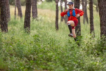 A woman in a red jacket is running through a forest. She is wearing a blue backpack and has her arms outstretched