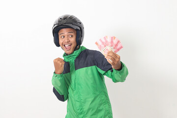 Portrait of Asian online taxi driver wearing green standing against white background, holding a bunch of rupiah money while raising fist, celebrating success