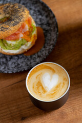 Heart latte art coffee scene with a bagel with egg, avocado, and fish on a wooden table. Perfect breakfast or brunch
