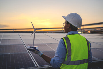 A man in a yellow vest is holding a wind turbine. The sky is orange and the man is wearing a hard...