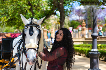 latin woman, brunette with curly hair, young and beautiful next to an elegant white horse pulling a...