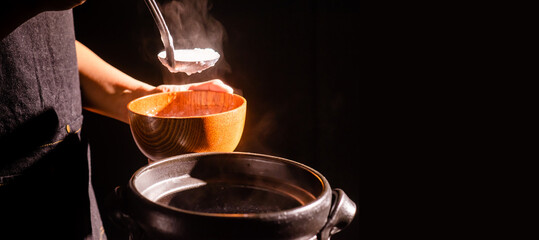 The cook was cooking rice porridge in a boiling clay pot with steam coming out and using a ladle to...