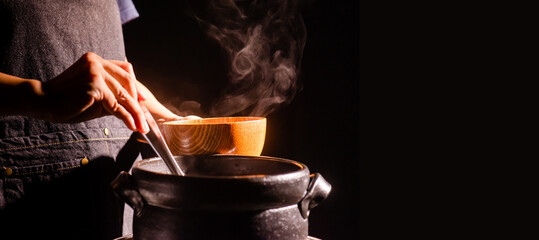 The cook was cooking in a boiling clay pot with steam coming out and using a ladle to scoop the food into a wooden bowl.