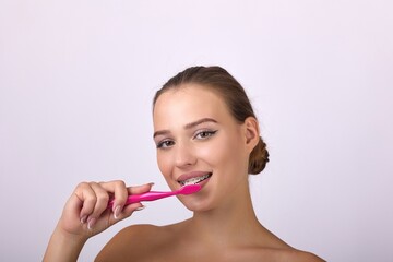 A young woman with a captivating smile brushing her teeth with a pink toothbrush. Her brown hair stands out against a white background, emphasizing her oral care routine.
