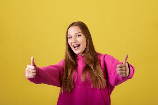 The image shows a young woman with long hair wearing a pink hoodie. She is smiling and showing thumbs up. The background is a bright yellow color. The image is taken in a studio and is well-lit.