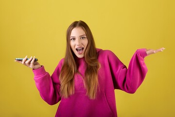 The image shows a young woman wearing a pink hoodie looking surprised while holding a phone in her...