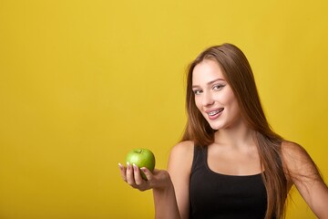 A young woman with braces on her teeth is smiling and holding a granny smith apple. She is wearing...