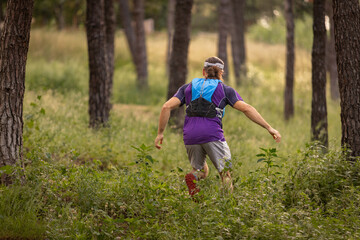 A man is running through a forest with a backpack on. The man is wearing a purple shirt and gray...