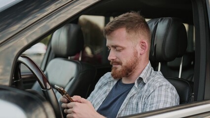 Smiling young man looking at smartphone screen, sitting in the car. Slow motion