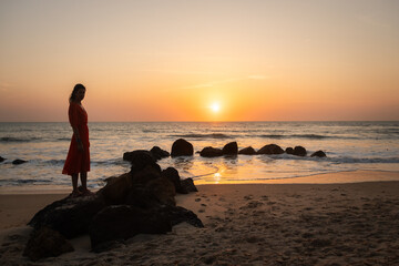 silhouette of woman at sunset standing in dress on rocks in the water on beach and ocean