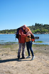 A man and a woman are hugging each other by a lake. They are carrying backpacks because they are hiking or trekking.